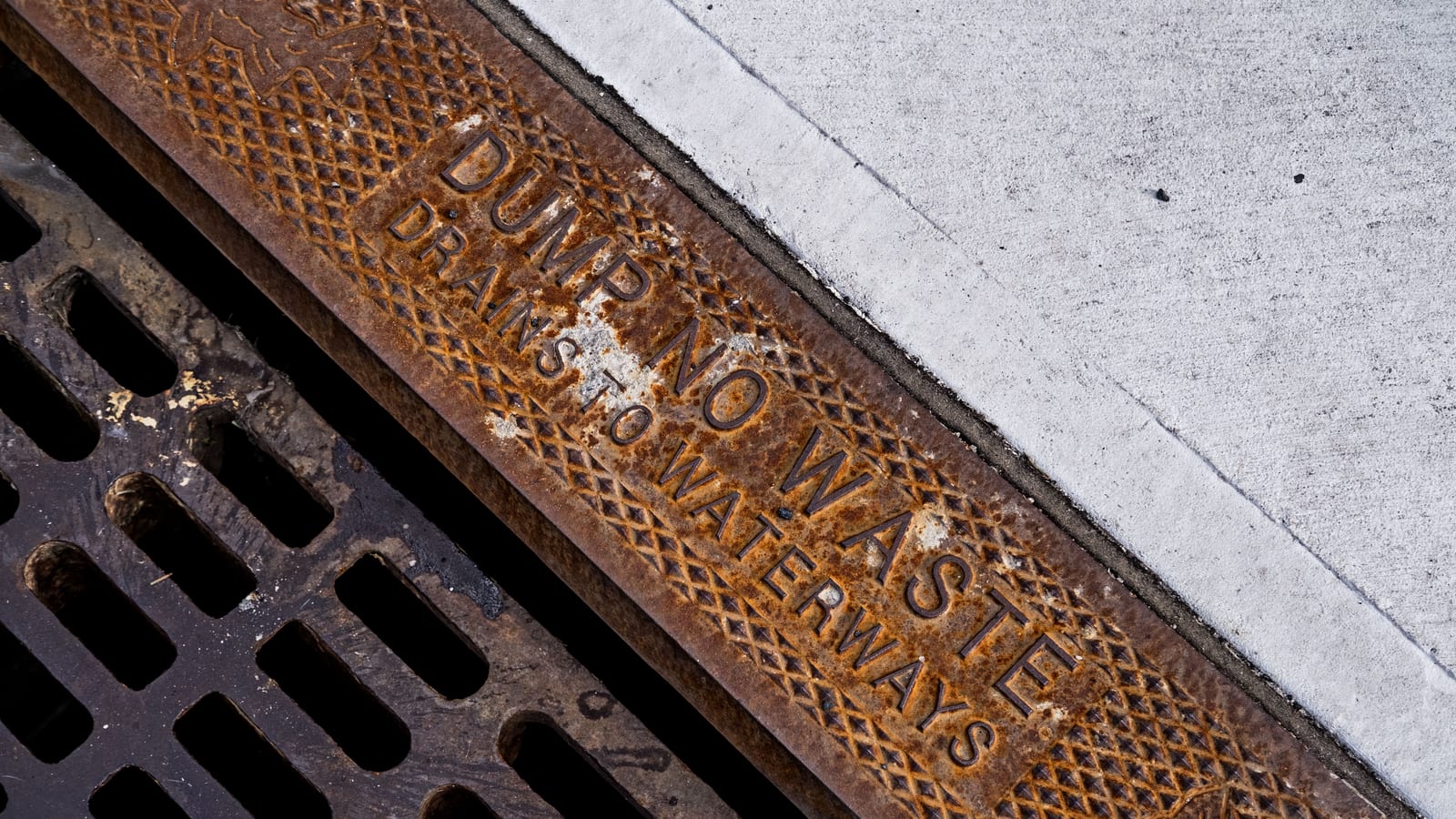 Close-up of a rusty drainage grate with environmental warning text on concrete sidewalk
