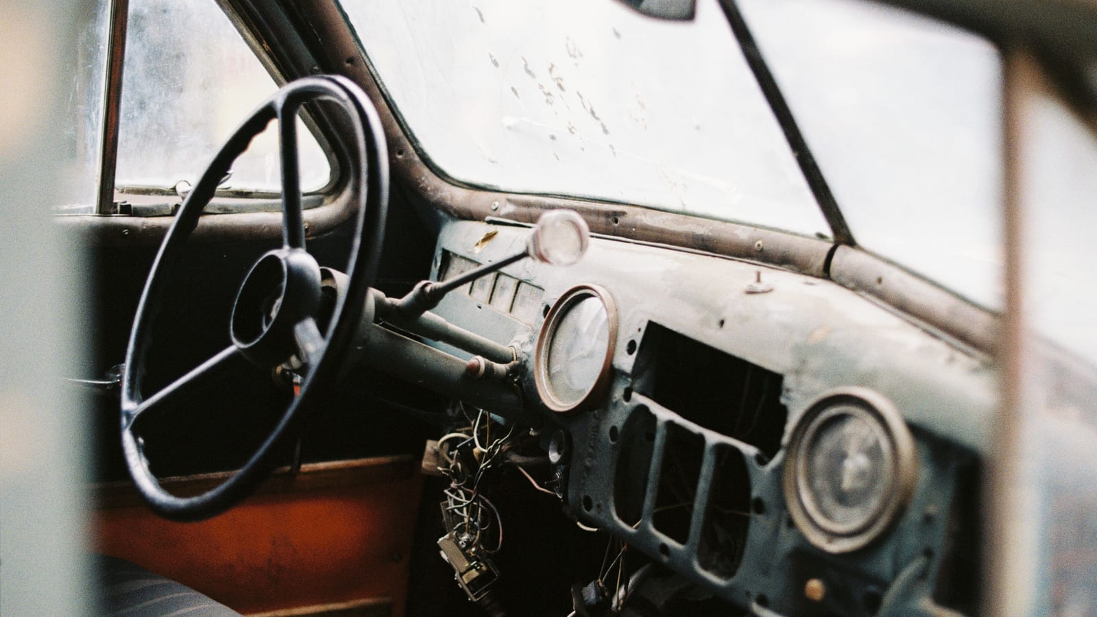 Interior of old fashioned automobile with seat and dirty windshield near big steering wheel and wire bundle above in daylight